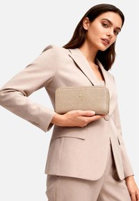 Portafoglio in pelle beige texturizzata con logo dorato, chiusura a zip e forma rettangolare. Presentato da una modella con un completo chiaro.