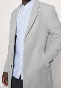 Mężczyzna w jasnoniebieskiej koszuli na guziki pod jasnoszarym wełnianym płaszczem, z ręką w kieszeni płaszcza na jednolitym tle.