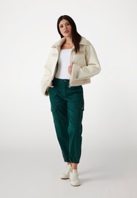 Fehér műszőrme dzseki magas gallérral, sötétzöld cargo nadrággal, amely zsebekkel rendelkezik. Fehér sportcipők teszik teljessé a megjelenést.