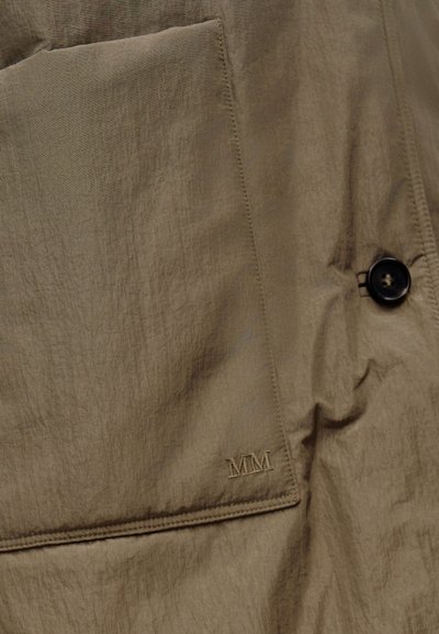 Barna szövetzseb közeli kép egy ruhadarabon, rajta hímzett "MM" logó és egy közeli fekete gomb.