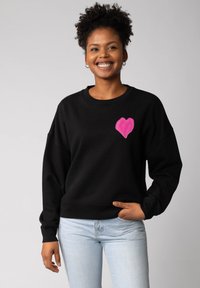 Glunderende vrouw met krullend haar, gekleed in een zwart sweatshirt met een roze hart op de borst en lichtblauwe jeans, staande tegen een effen achtergrond.