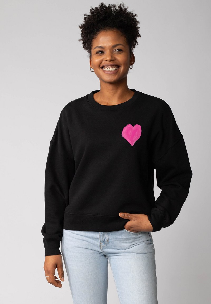 Glunderende vrouw met krullend haar, gekleed in een zwart sweatshirt met een roze hart op de borst en lichtblauwe jeans, staande tegen een effen achtergrond.