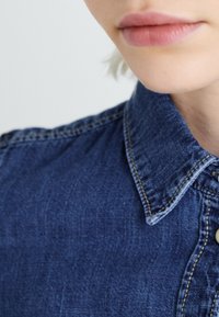 Blue denim shirt featuring a buttoned collar, visible stitching details, and a structured fit. Close-up of collar area and fabric texture.