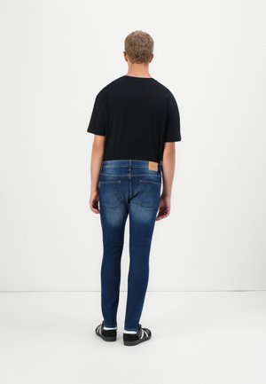 Homme portant un t-shirt noir, un jean skinny bleu et des baskets noires, debout face à un mur blanc, les mains détendues le long du corps.