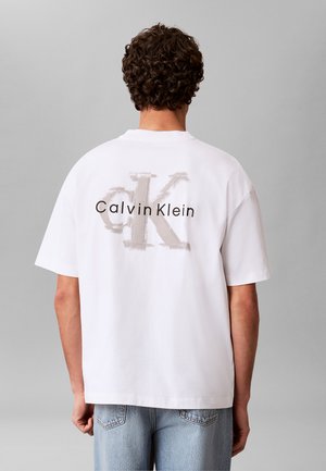 Mand med krøllet hår iført en hvid Calvin Klein t-shirt med stort "ck" logo og tekst på bagsiden, kombineret med lyseblå jeans.