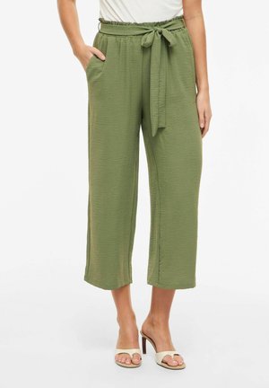 Olivne zelene kratke široke hlače z zavezovanjem v pasu, kombinirane s bež sandalami z odprtimi prsti in peto ter v belo vpojeno zgornjo majico.