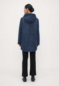 Kvinna med mörkt hår i en knut, iförd en marinblå hooded regnkappa, svarta byxor och svarta höga klackar, stående mot en enkel ljus bakgrund, vänd bort.