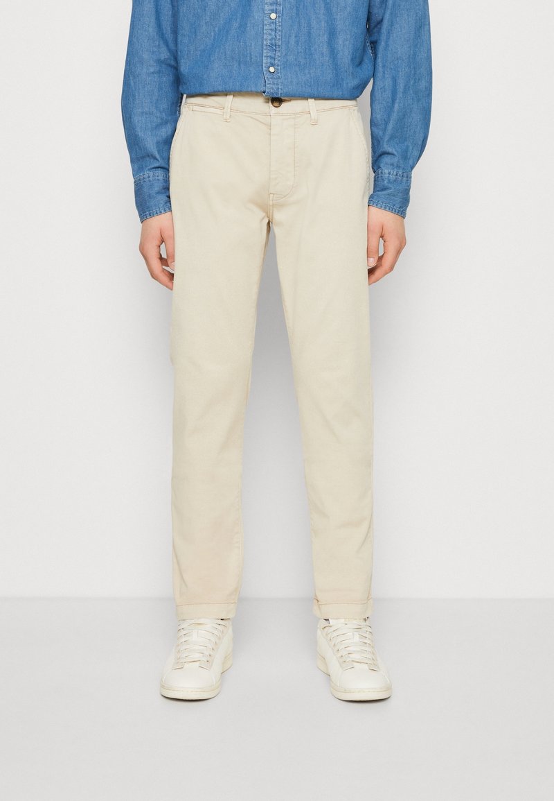 Beige cotton trousers with a straight cut, combined with a blue chambray button-up shirt and white sneakers.