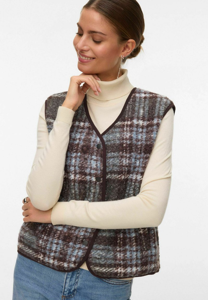 Femme souriante, main près du menton, portant un col roulé crème, un jean bleu et un gilet à carreaux boutonné aux tons marron et bleu.