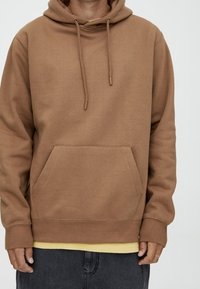 Sudadera con capucha marrón hecha de tela suave con un bolsillo canguro, capucha con cordón, puños y dobladillo acanalados. Lleva una camiseta amarilla debajo.