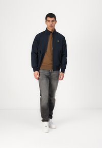 Giubbotto bomber navy con collo rialzato, caratterizzato da un logo giallo sul petto, abbinato a un maglione marrone e jeans grigi sbiaditi. Sneakers bianche.