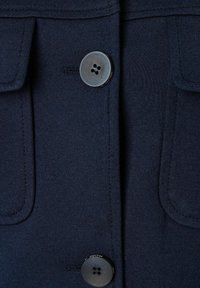 Navyblauwe stof met een gladde textuur, met twee zakken en een grote, ronde zwarte knoopdetail aan de voorkant.
