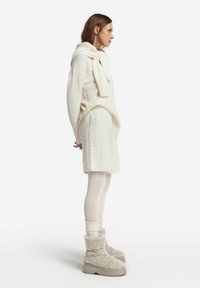 Chunkig krämfärgad stickad tröja med huva, ihop med en kabelstickad krämfärgad kjol, krämfärgade leggings och fluffiga beige ankelstövlar. Texturerade detaljer synliga.