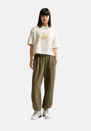 Mujer de pie con las manos en los bolsillos, vistiendo una camiseta corta blanca de Nike, pantalones deportivos verde oliva y zapatillas blancas Nike sobre fondo blanco.