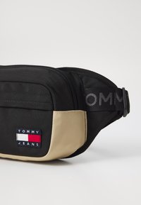Banane noire et beige en tissu résistant, dotée d'une poche zippée, d'un patch logo et d'une sangle ajustable avec le texte "TOMMY".