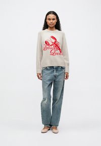 Mujer con cabello rizado vestida con un suéter beige con un langosta roja y el texto "Amante de las Langostas", jeans rotos y holgados, y zapatos planos beige, de pie frente a un fondo blanco.