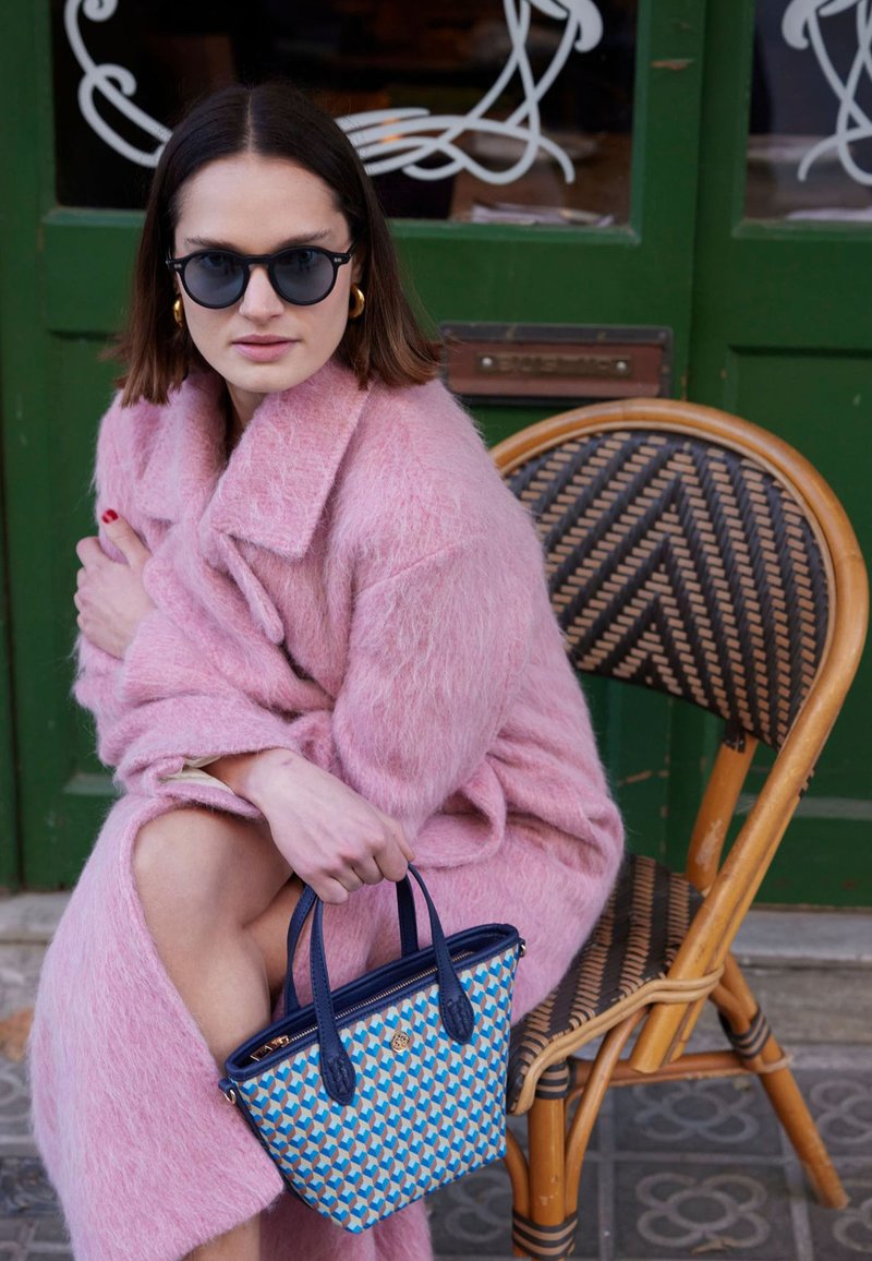 Pink, fuzzy oversized coat; blue patterned handbag with dark handles; round black sunglasses; rattan chair; green background.