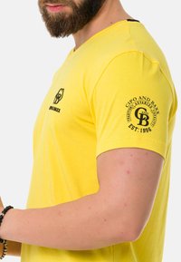 Gelbes Kurzarm-T-Shirt aus weichem Stoff, mit V-Ausschnitt und schwarzen Drucklogos auf der Brust und dem Ärmel.
