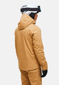 Giacca da sci in tessuto marrone chiaro, con cappuccio, tasche laterali e polsini elasticizzati. Indossata con casco bianco e guanti neri.