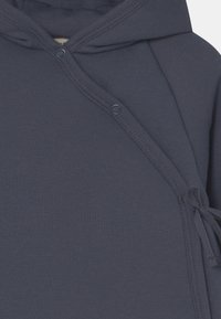 Haut à capuche bleu marine en tissu doux et texturé, doté d'une fermeture à pression à l'avant et d'un accent noué à l'ourlet pour un ajustement ajustable.
