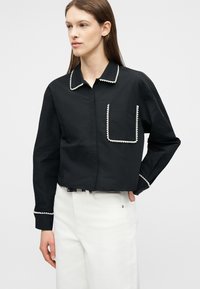 Mujer con camiseta negra de manga larga con ribete de perlas blancas en el cuello, el bolsillo y los puños, combinada con pantalones blancos contra un fondo liso.