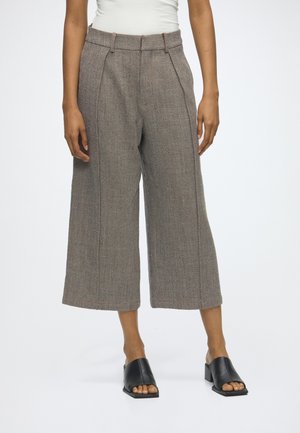 Person iført højtaljede, bredbenede cropped brune teksturerede bukser, hvid top og sorte åbne hælede mule, stående på en ensfarvet baggrund.