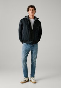 Chaqueta bomber negra con capucha gris, llevada sobre una camiseta blanca. Combinada con unos vaqueros de mezclilla claros y desgastados y zapatillas beige con detalles en negro.