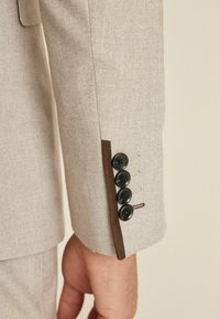 Bežani herringbone sako sa rukavima koji sadrži pet crnih dugmadi, kontrastnim smešnim detaljima i vidljivim šavovima.