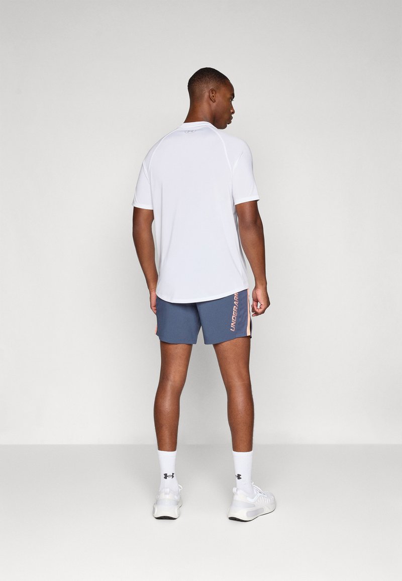 Weißes, kurzärmliges Sportshirt kombiniert mit dunkelgrauen Shorts mit pinken Akzenten. Das Modell steht mit dem Rücken zur Kamera, trägt weiße Turnschuhe und Socken.