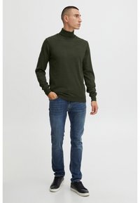 Hombre con un suéter de cuello alto verde oscuro, jeans azules y zapatillas negras, de pie con una mano en el bolsillo, mirando a la derecha.