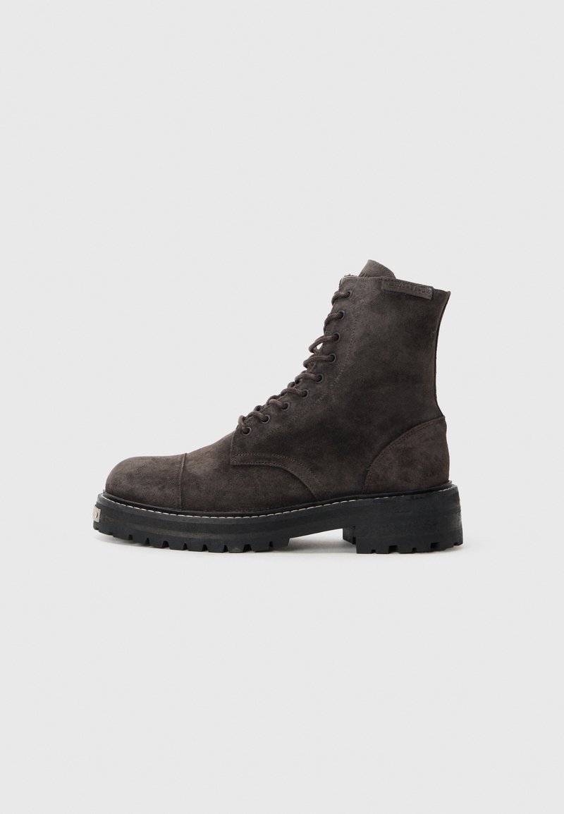 Stivaletto alla caviglia in suede grigio scuro con punta rotonda, chiusura con lacci e suola in gomma nera spessa con grip testurizzato. Dettagli di design minimalista.