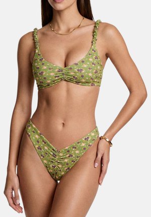Donna che indossa un bikini verde floreale arricciato con spalline arricciate, collana e braccialetto in oro, in piedi davanti a uno sfondo semplice.