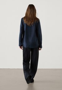 Camicia lunga in satin blu navy con orlo arrotondato, abbinata a pantaloni larghi neri, posizionata su uno sfondo chiaro e semplice.