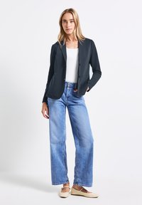 Tmavě modrý blazer se dvěma předními kapsami, nošený přes bílý top, spárovaný s světlomodrými širokými džíny a béžovými plochými botami. Hladký, přiléhavý design.