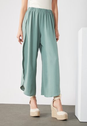 Mujer con pantalones anchos de pierna en verde claro y alpargatas cuña beige con correas en el tobillo, de pie sobre suelo gris frente a fondo blanco.