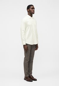 Camisa creme clara com botões, corte ajustado, combinada com calças castanhas texturizadas e sapatos de couro castanhos. Gola simples e mangas compridas.