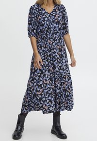 Květinové midi šaty s černým pozadím, se modrými a růžovými květy, nabíranými rukávy, výstřihem do V a nařaseným pasem, doplněné černými kotníkovými botami.