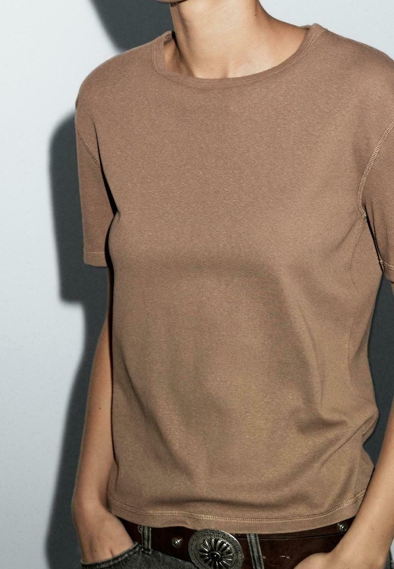 Personne portant une chemise marron unie à manches courtes rentrée dans un jean avec une ceinture foncée et une boucle argentée, debout avec une main dans la poche.