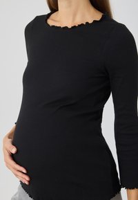 Black ribbed long-sleeve top with a scalloped neckline, fitted design, and visible stretch for accommodating a pregnant shape.