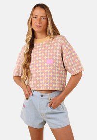 Pull court avec un motif en losanges jaune et lavande, comportant un petit écusson logo rose. Associé à un short en denim clair.
