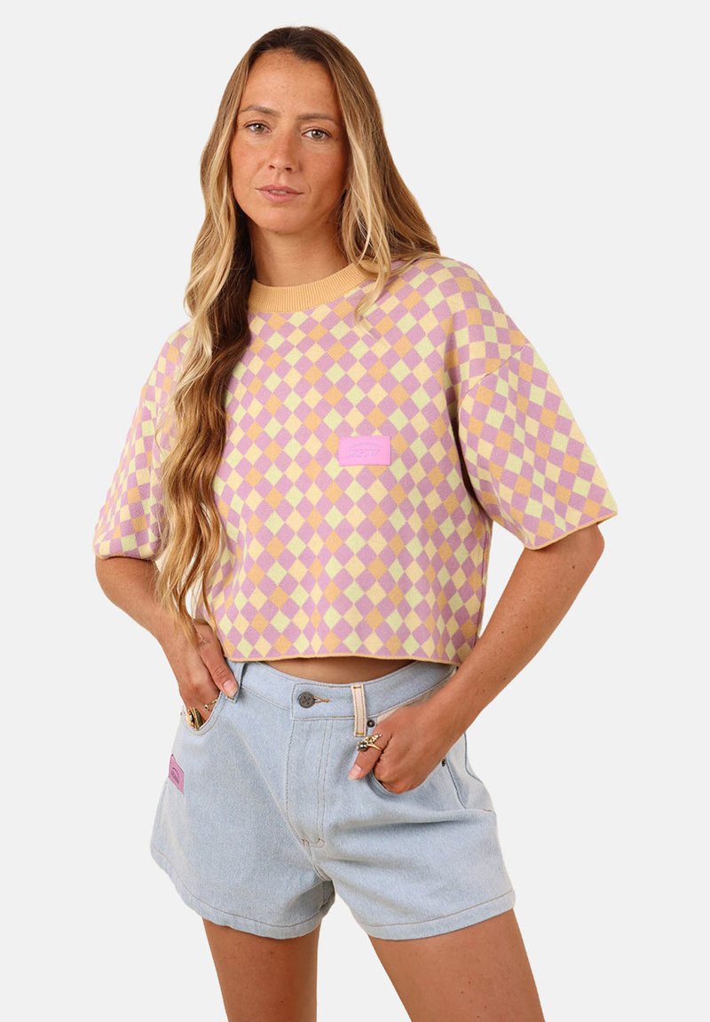 Pull court avec un motif en losanges jaune et lavande, comportant un petit écusson logo rose. Associé à un short en denim clair.