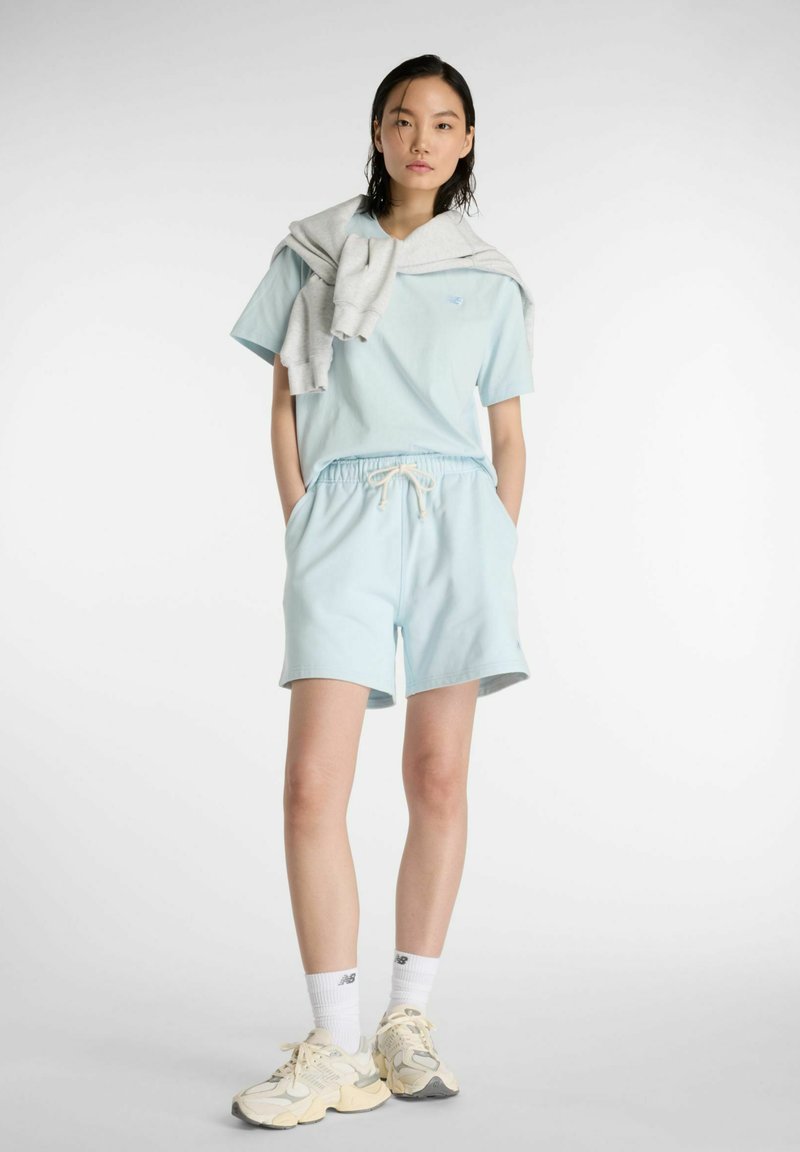 Maglietta di cotone azzurro e pantaloncini abbinati, con coulisse regolabile. Maglione grigio appoggiato sulle spalle. Scarpe da ginnastica.