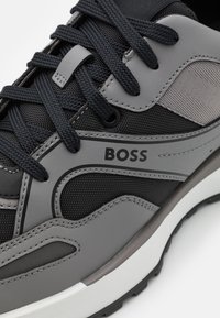 Chaussure de sport grise et noire avec un maillage texturé et des panneaux en cuir lisse, des lacets ronds, et une semelle blanche avec des accents subtils.