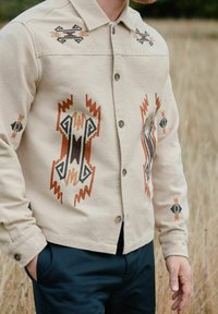 Beige overhemd met knoopsluiting, versierd met geometrische borduursels in zwart, oranje en bruin. Het heeft een kraag en lange mouwen met knoopmanchet.