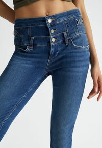 Jeans in denim blu a vita alta presentano chiusure frontali con bottoni, cinque tasche e una finitura leggermente sbiadita, con una texture liscia e lucida.