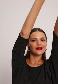 Czarna błyszcząca bluzka z długim rękawem, luźny krój i szeroki dekolt. Modelka trzyma ręce nad głową, prezentując złote kolczyki w kształcie łzy i wyrazisty czerwony błyszczyk do ust.
