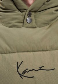 Olivgrüne Kapuzenjacke mit strukturierter Oberfläche, ausgestattet mit zwei metallischen Knöpfen und einem schwarzen gestickten Logo auf der Brust.