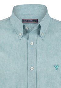 Camisa button-up de un azul claro con una textura sutil, un cuello puntiagudo y un pequeño logo de ave bordado en el pecho.