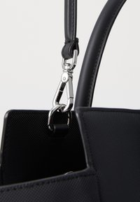 Sac à main noir avec un extérieur texturé, une sangle en cuir et un clip en métal argenté pour attacher, mettant en avant des éléments de design moderne.