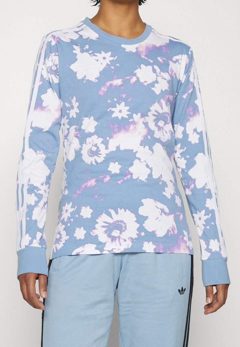 Ljusblå långärmad skjorta med blommönster i vitt och lila. Tre vita ränder på varje ärm. Rund halsringning och mjuk textur.